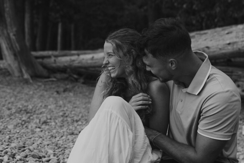 Black and white image of engaged couple seated together on the pebbled Lake McDonald beach, woman laughing with her head thrown back as her partner leans in close, driftwood and cedar trees behind them — Ryan + Liz — Surprise Proposal — Glacier National Park — Stacy Edwards Photography