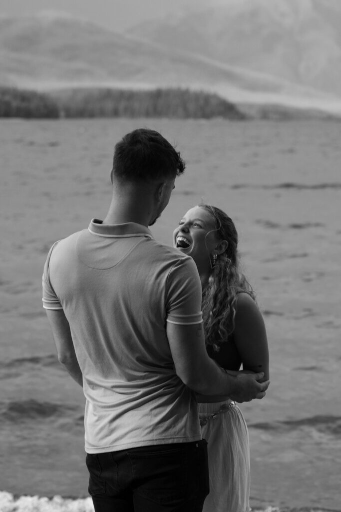 Black and white photo of newly engaged couple laughing together at the water's edge, Glacier National Park — Ryan + Liz — Surprise Proposal — Glacier National Park — Stacy Edwards Photography