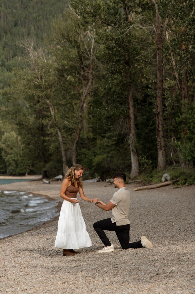 Man on one knee proposing to woman on the pebbled beach at Apgar Village, Lake McDonald — Ryan + Liz — Surprise Proposal — Glacier National Park — Stacy Edwards Photography