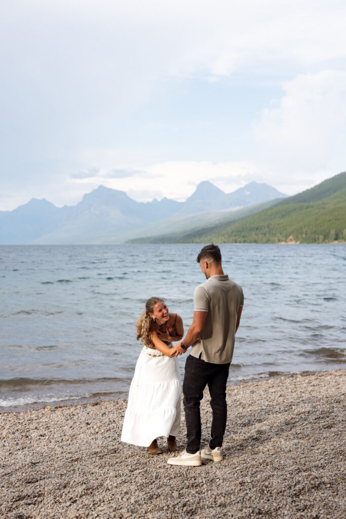 Newly engaged couple holding hands and smiling on the shores of Lake McDonald with Glacier peaks in the background — Ryan + Liz — Surprise Proposal — Glacier National Park — Stacy Edwards Photography