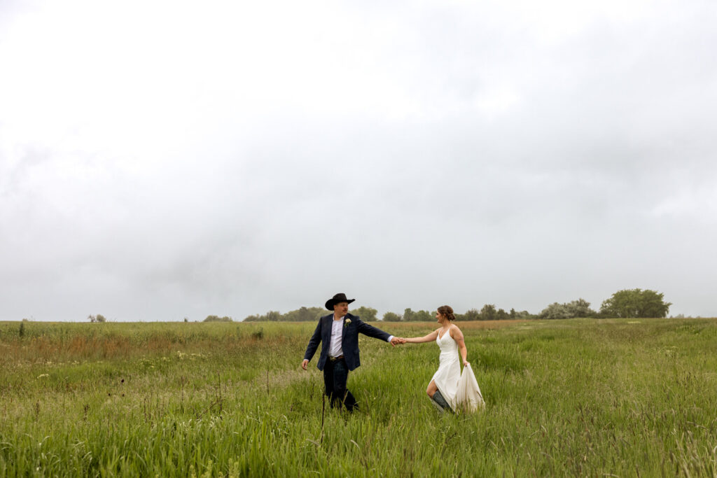 Wide shot of bride and groom holding hands, walking through a grassy field — Aislynn + Kyle — Mission Valley Wedding — Charlo Montana — Stacy Edwards Photography