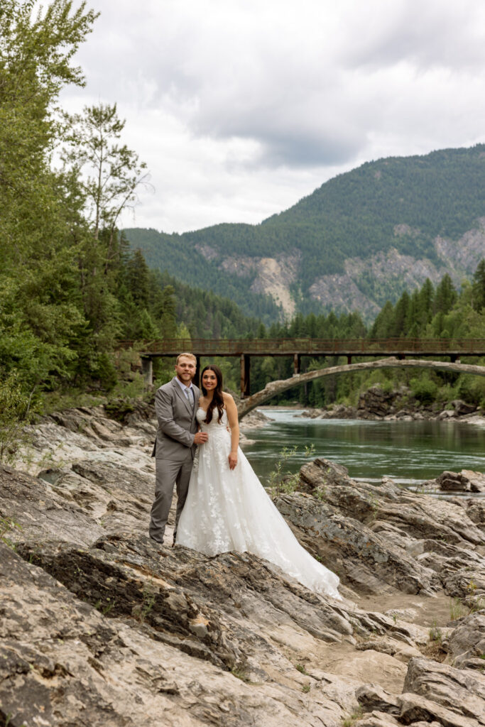 Bride and groom portrait on the rocky shore of Belton Bridge with the Middle Fork Flathead River and mountains behind them — Glacier National Park elopement photographer Stacy Edwards Photography