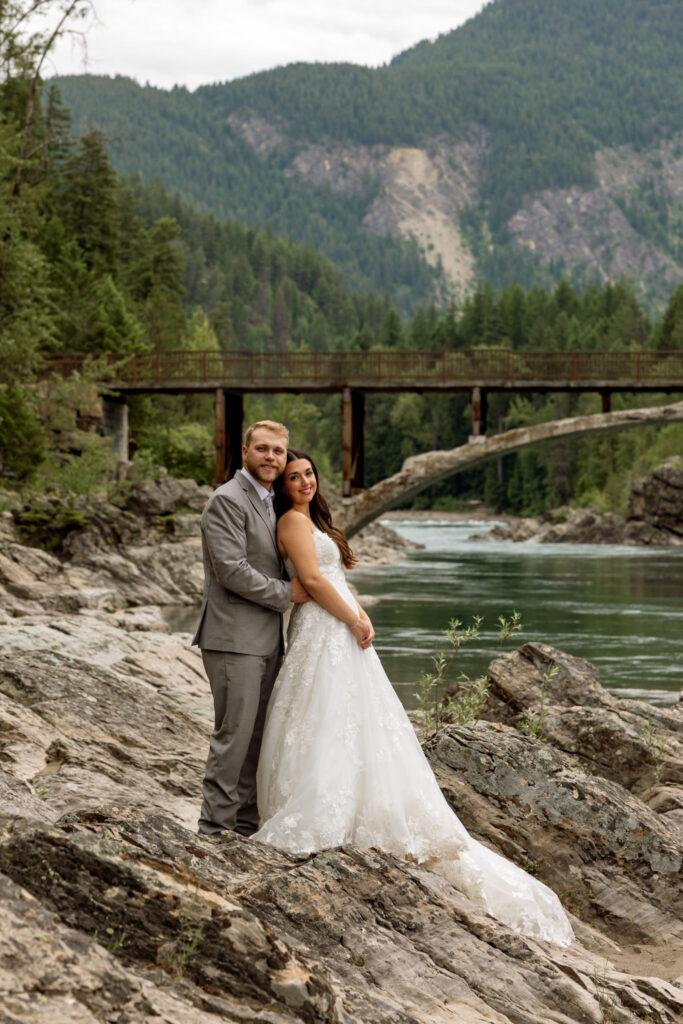 Bride and groom embracing on the rocky shoreline at Belton Bridge with the river and mountains in the background — Glacier National Park wedding photographer Stacy Edwards Photography