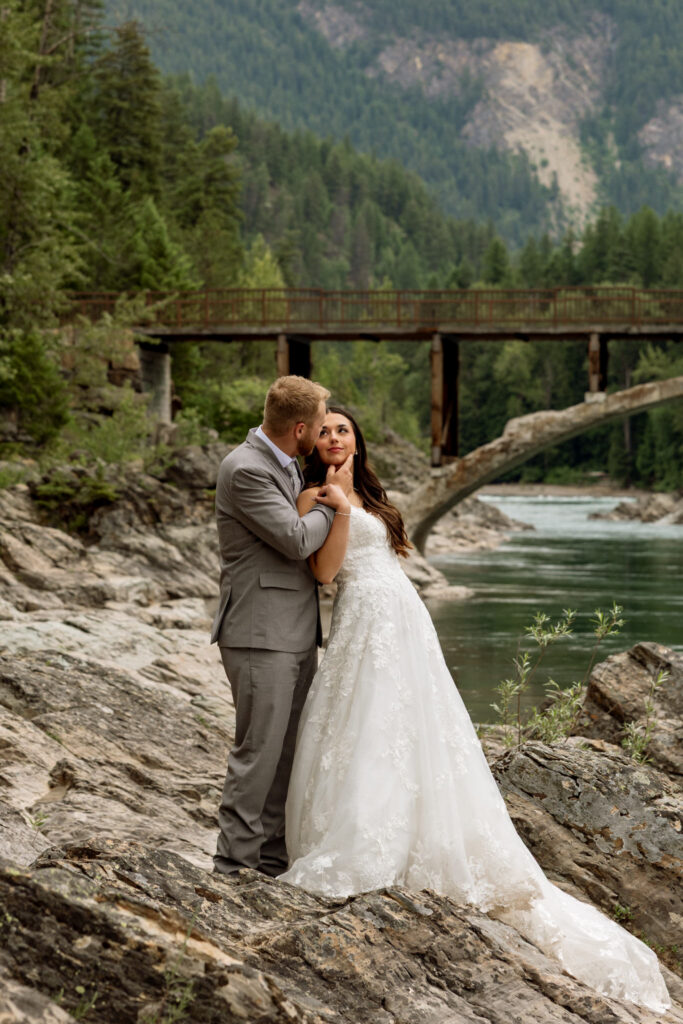 Groom kissing bride on the cheek at Belton Bridge with the Middle Fork Flathead River behind them — Glacier National Park elopement photographer Stacy Edwards Photography