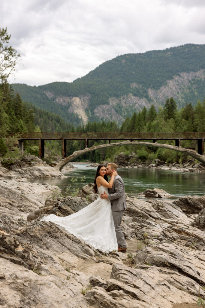 Bride and groom kissing on the rocky shore at Belton Bridge with wedding dress train spread across the rocks — West Glacier Montana elopement — Stacy Edwards Photography
