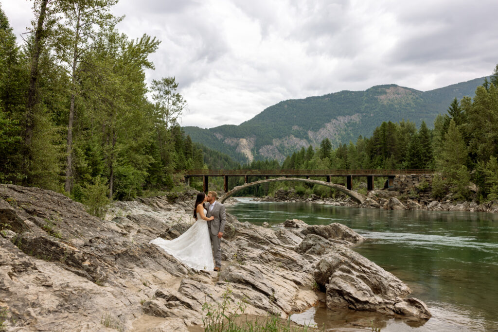 Bride and groom kissing at Belton Bridge with the river and forested mountains in the background — Glacier National Park elopement photographer Stacy Edwards Photography