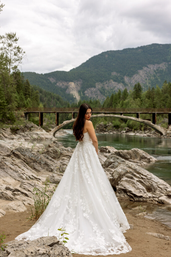 Bride in lace wedding gown standing on the rocks at Belton Bridge with back to camera — West Glacier Montana elopement — Stacy Edwards Photography