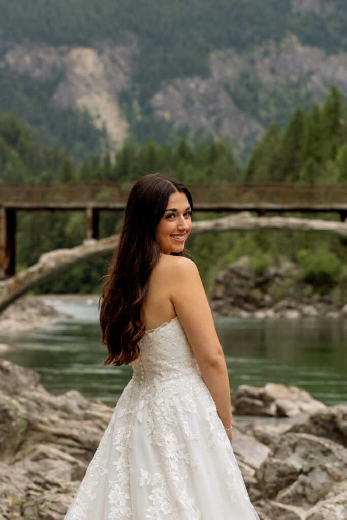 Bride looking over her shoulder and smiling on the rocky shore at Belton Bridge — Glacier National Park elopement — Stacy Edwards Photography