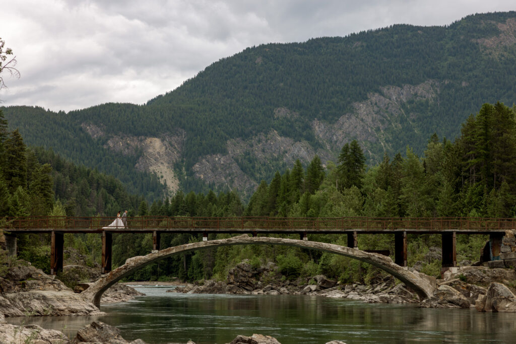 Wide shot of bride and groom standing together on Belton Bridge over the Middle Fork Flathead River — West Glacier Montana elopement — Stacy Edwards Photography