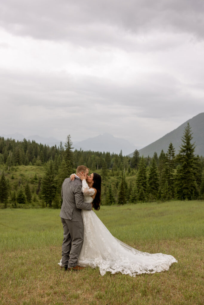 Bride and groom kissing in a green meadow with mountain peaks and pine trees behind them — Belton Stage Park Glacier National Park elopement — Stacy Edwards Photography