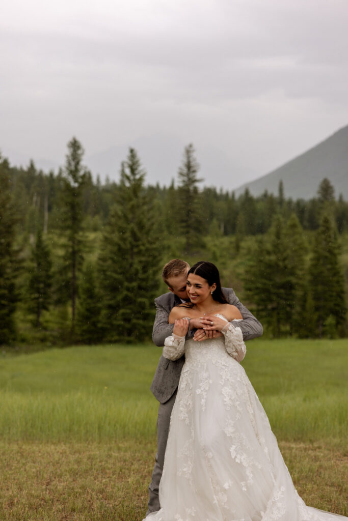 Groom kissing bride on the cheek from behind in a rainy green meadow near Glacier National Park — Glacier National Park elopement — Stacy Edwards Photography