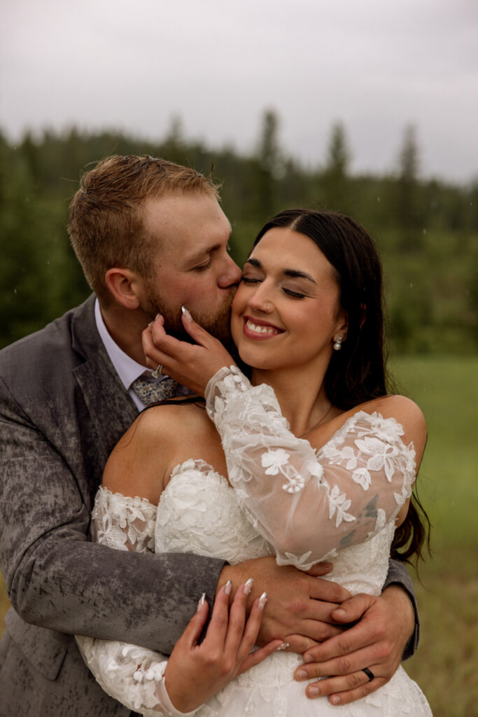 Groom kissing bride on the cheek while holding her close in a meadow near Glacier National Park — Belton Stage Park elopement — Stacy Edwards Photography