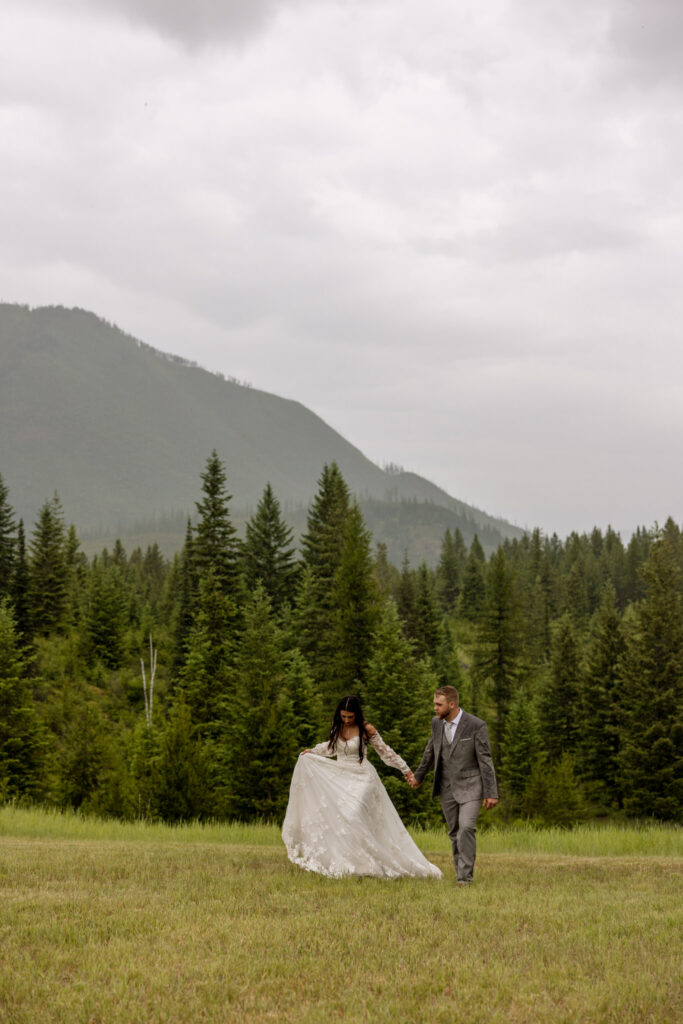 Bride and groom walking hand in hand through a meadow with pine trees and mountain peak behind them — Belton Stage Park Glacier National Park elopement — Stacy Edwards Photography