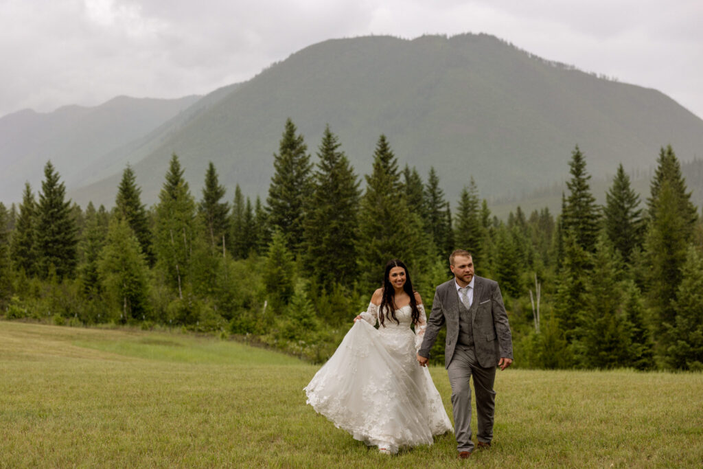 Bride and groom walking hand in hand through a green meadow with a dramatic mountain peak behind them — Glacier National Park elopement photographer Stacy Edwards Photography