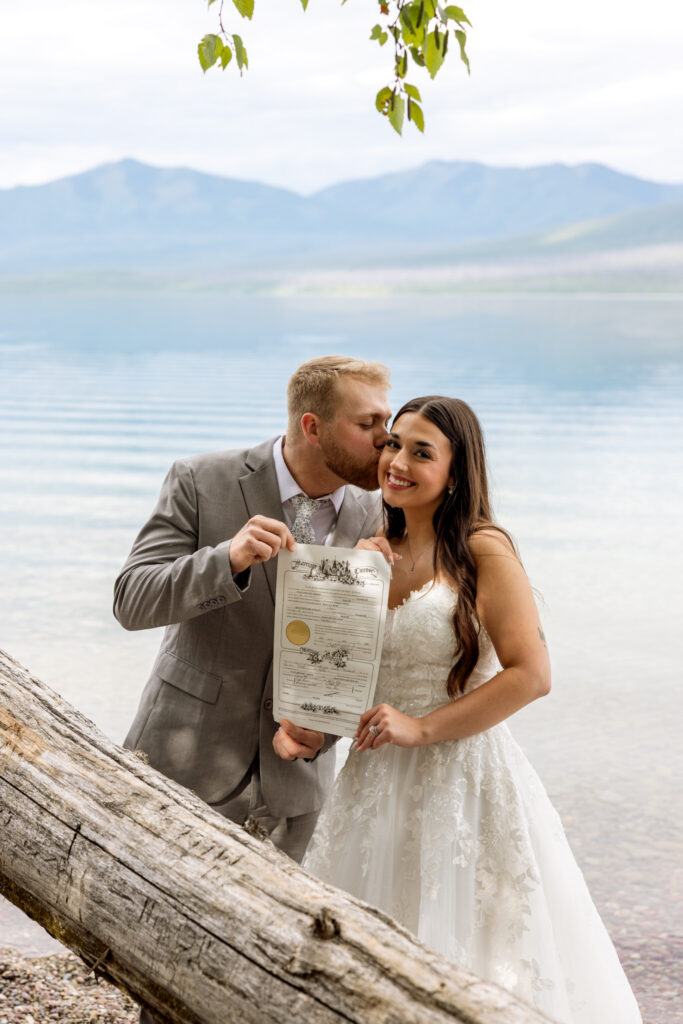 Bride and groom holding signed marriage license on the shore of Lake McDonald after elopement ceremony — Jackson Bay Glacier National Park — Stacy Edwards Photography