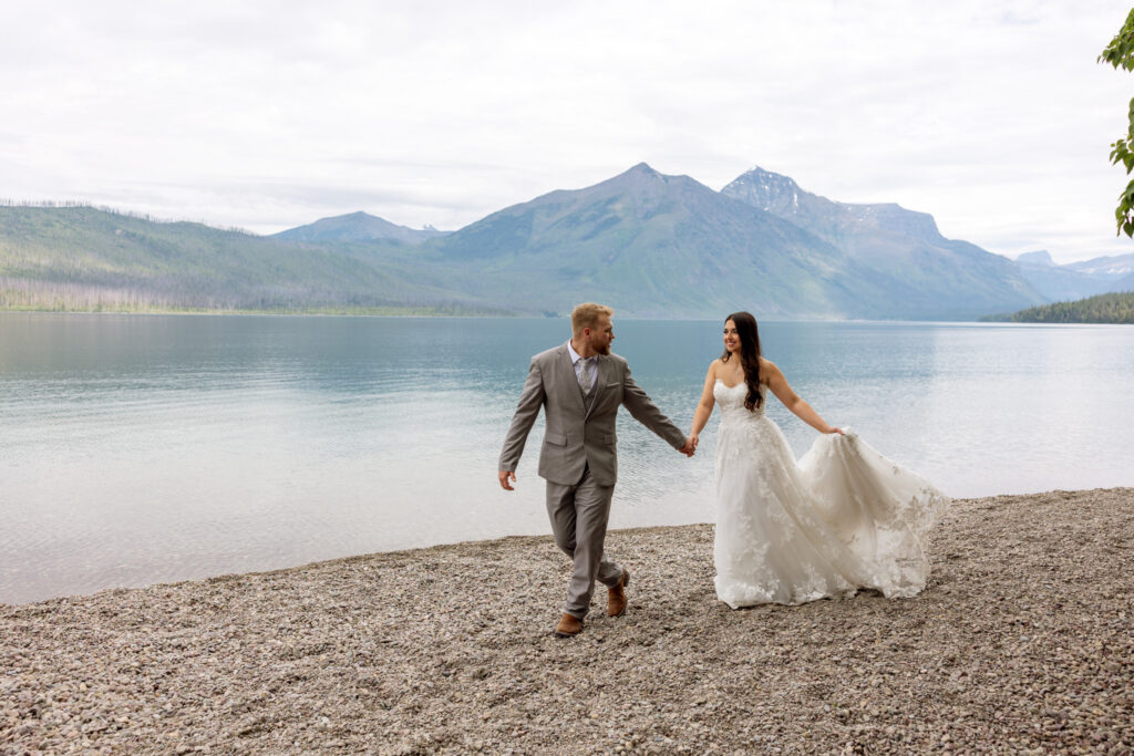 Bride and groom walking hand in hand along the shore of Lake McDonald with mountain peaks behind them — Glacier National Park wedding photographer Stacy Edwards Photography