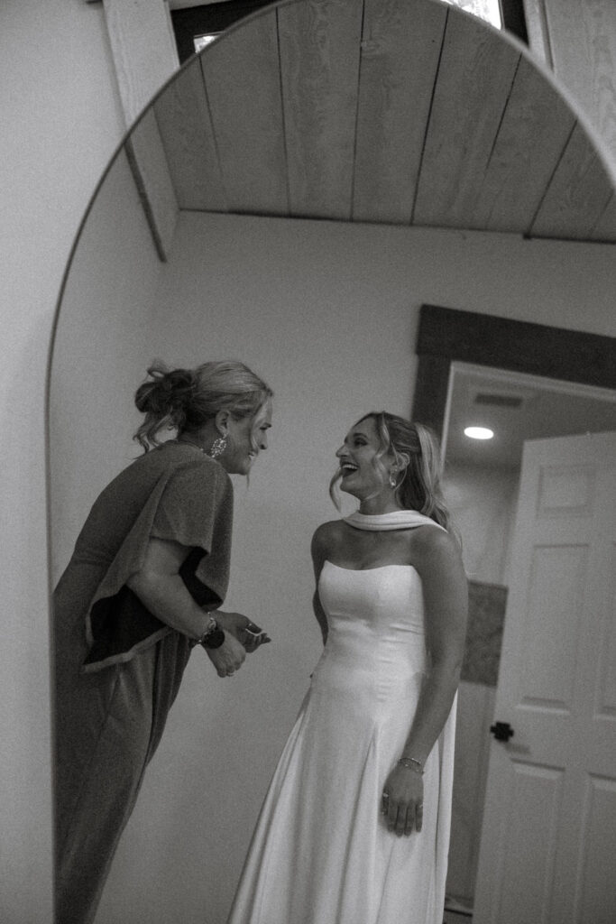 Black and white photo of bride laughing with mother while looking in arched mirror before ceremony — Micah + Greg — Ember & Altar Wedding — Polson Montana — Stacy Edwards Photography