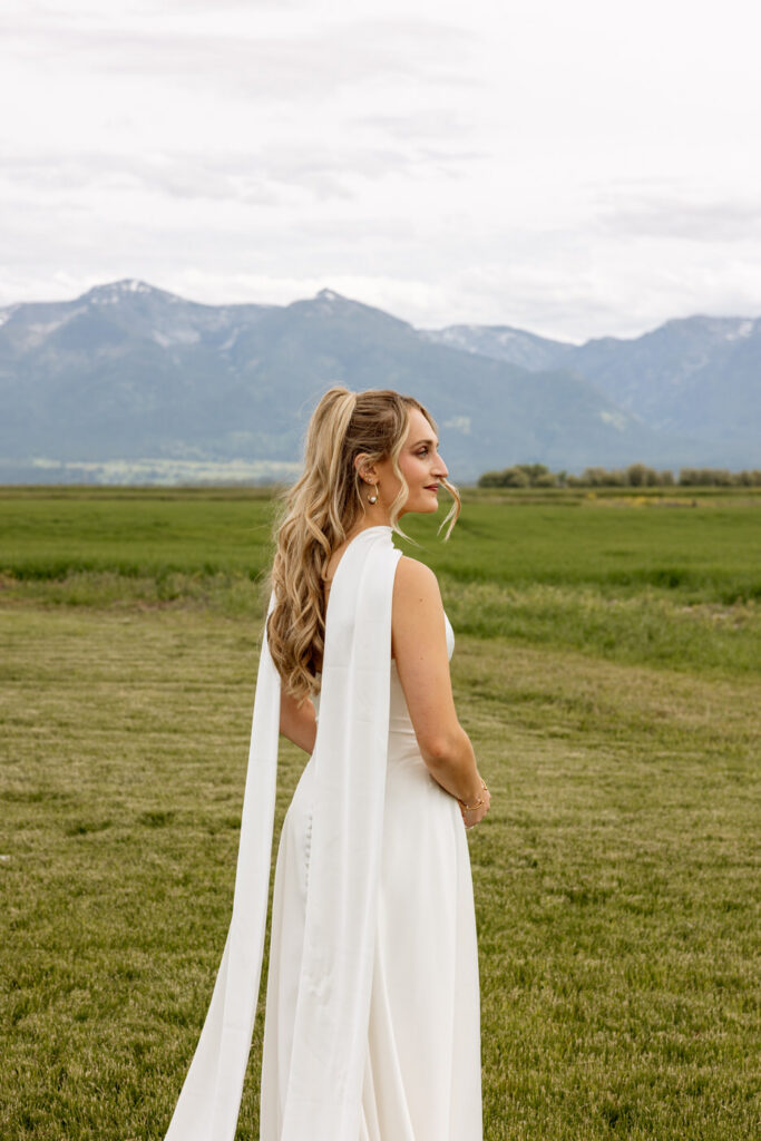 Bride in sleek white gown with flowing scarf over her shoulder toward Mission Mountains — Micah + Greg — Ember & Altar Wedding — Polson Montana — Stacy Edwards Photography
