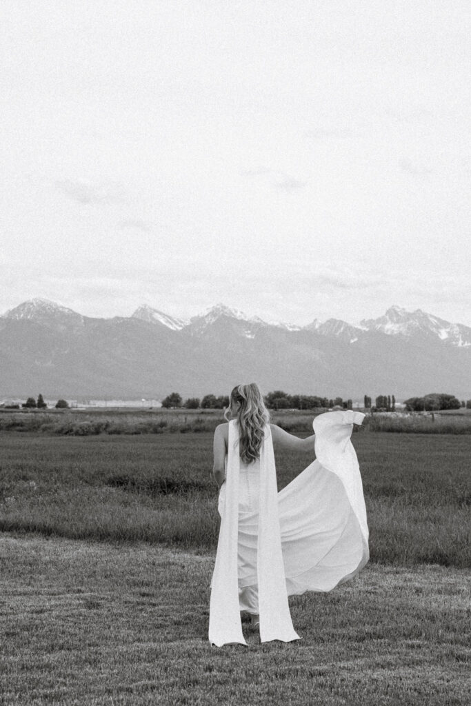Black and white photo of bride facing away holding dress out in green field with Mission Mountains behind her — Micah + Greg — Ember & Altar Wedding — Polson Montana — Stacy Edwards Photography