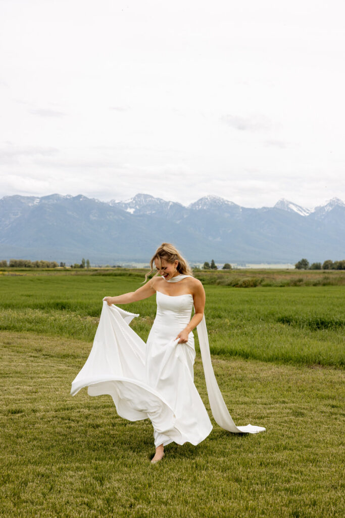 Bride laughing and twirling dress in open green field with snow-capped Mission Mountains behind her — Micah + Greg — Ember & Altar Wedding — Polson Montana — Stacy Edwards Photography