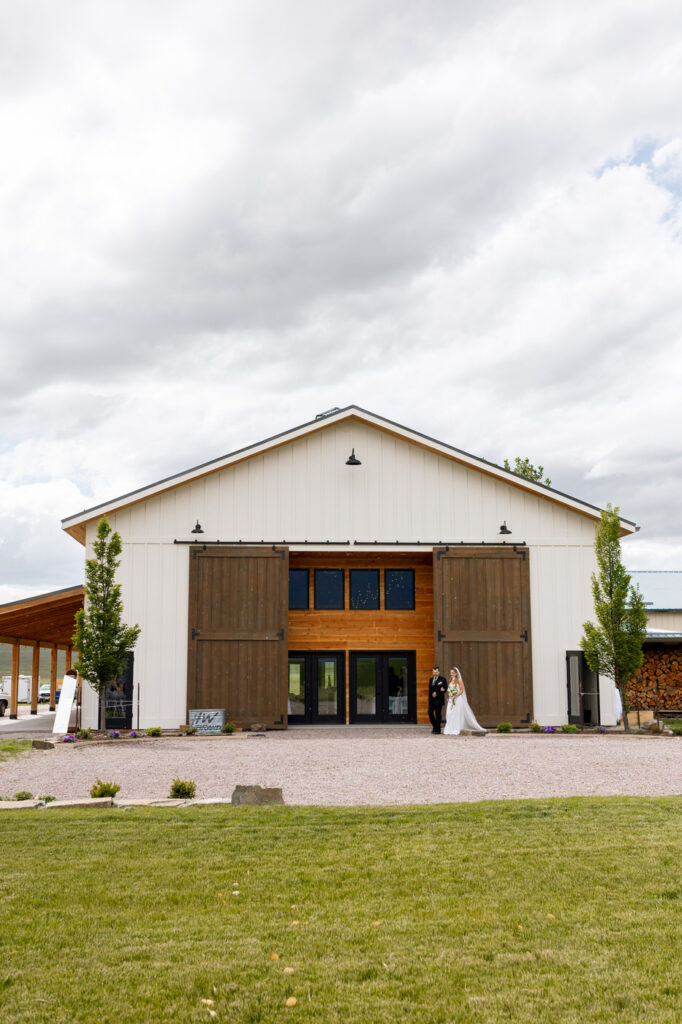 Wide shot of Ember and Altar barn exterior with bride and groom walking out through large sliding barn doors — Micah + Greg — Ember & Altar Wedding — Polson Montana — Stacy Edwards Photography
