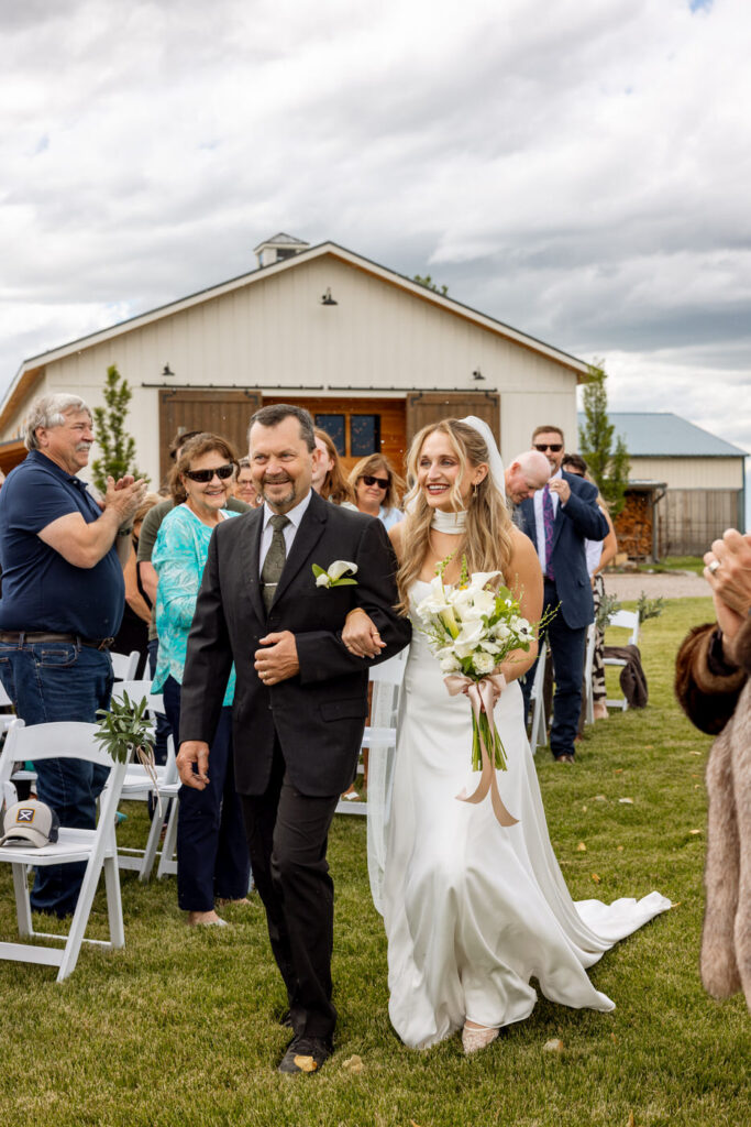 Bride walking down the aisle on father's arm with Ember and Altar barn in the background — Micah + Greg — Ember & Altar Wedding — Polson Montana — Stacy Edwards Photography