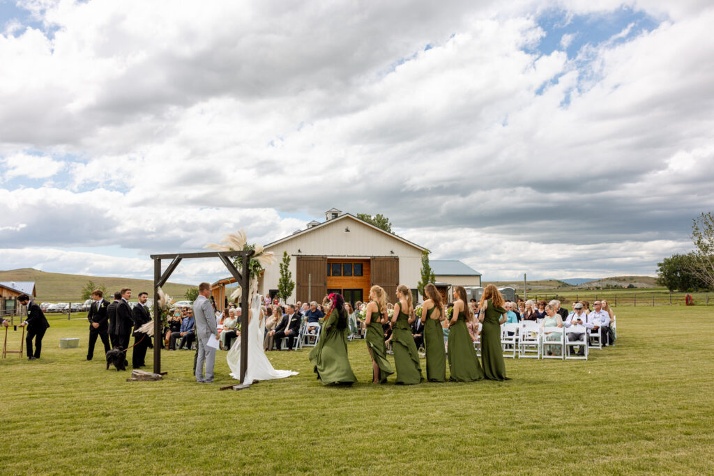 Wide shot of outdoor ceremony with wedding party at dark wood arch, guests seated on white chairs, and Ember and Altar barn behind — Micah + Greg — Ember & Altar Wedding — Polson Montana — Stacy Edwards Photography