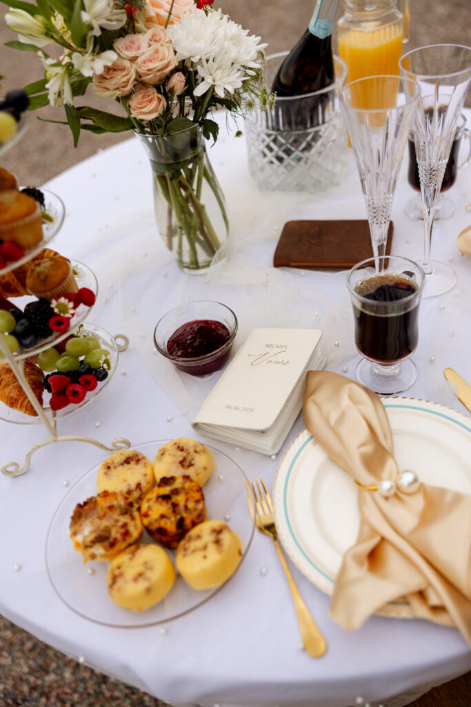 Close-up of outdoor sweetheart table with vow booklet, egg bites, fresh fruit tower, and mimosa setup — Micah + Greg — Ember & Altar Wedding — Polson Montana — Stacy Edwards Photography
