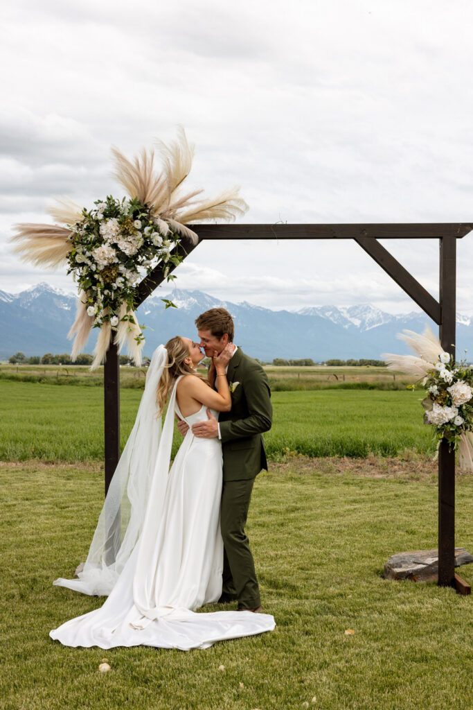 Bride and groom kissing at outdoor altar with pampas grass arch and green Mission Valley fields stretching behind them — Micah + Greg — Ember & Altar Wedding — Polson Montana — Stacy Edwards Photography