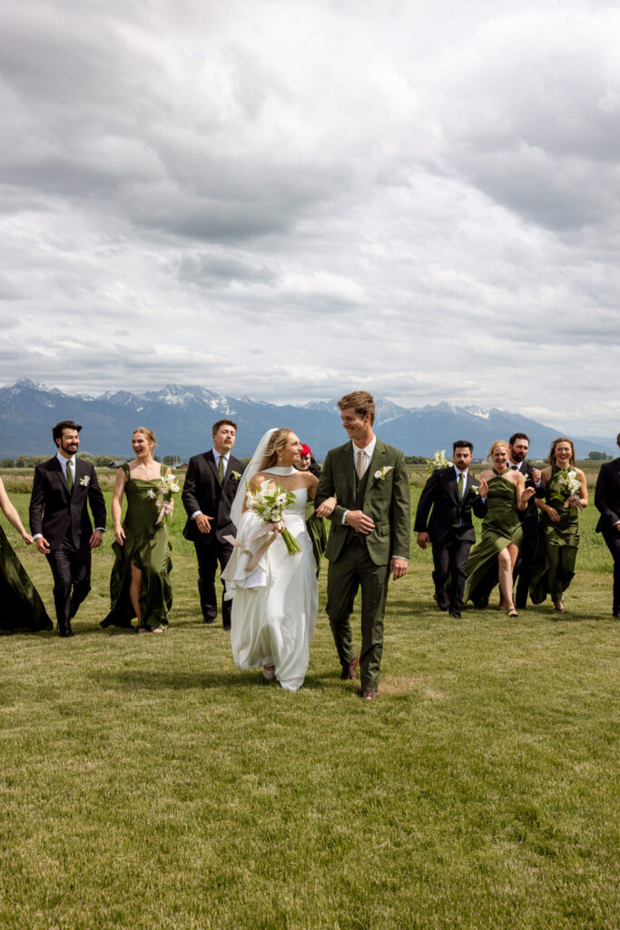 Bride and groom leading the wedding party walking through open field with Mission Mountains in the background — Micah + Greg — Ember & Altar Wedding — Polson Montana — Stacy Edwards Photography