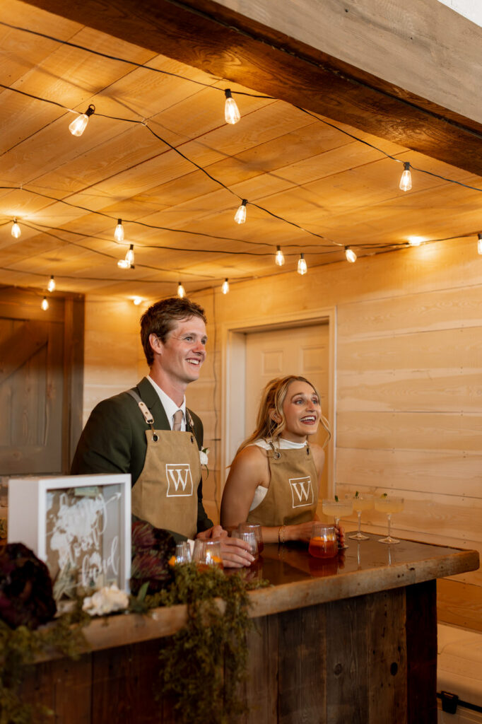 Bride and groom wearing monogrammed aprons laughing together behind the cocktail hour bar under Edison string lights — Micah + Greg — Ember & Altar Wedding — Polson Montana — Stacy Edwards Photography