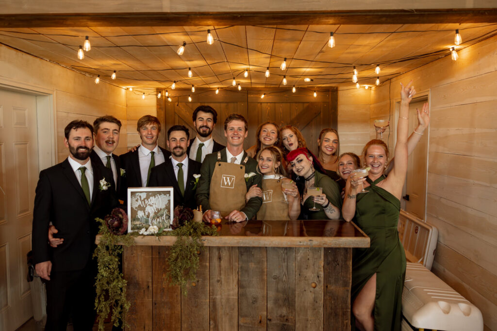 Bride and groom with full wedding party gathered and celebrating behind the cocktail bar under Edison string lights — Micah + Greg — Ember & Altar Wedding — Polson Montana — Stacy Edwards Photography