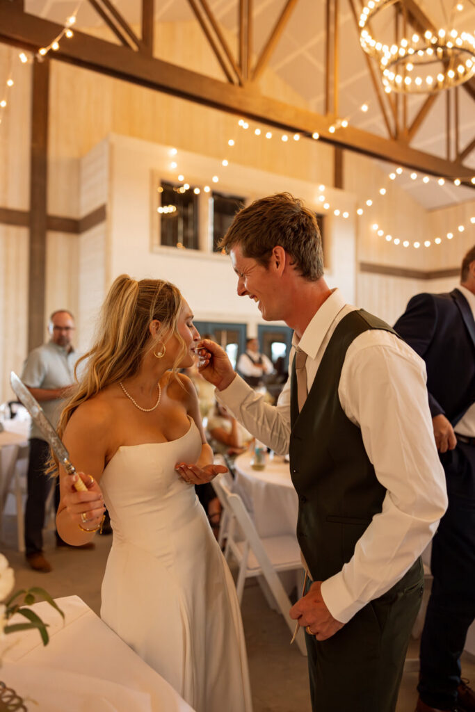 Groom gently feeding bride a bite of wedding cake while she laughs inside the reception hall — Micah + Greg — Ember & Altar Wedding — Polson Montana — Stacy Edwards Photography