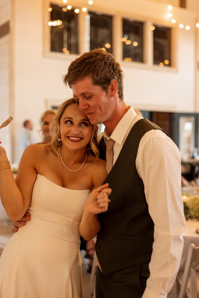 Groom kissing bride on the temple during reception while she smiles up at him under warm string lights — Micah + Greg — Ember & Altar Wedding — Polson Montana — Stacy Edwards Photography