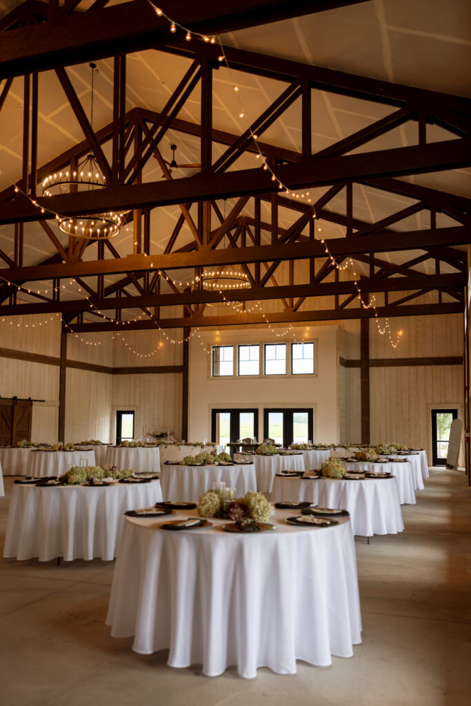 Reception hall interior with vaulted wood truss ceiling, globe string lights, ring chandeliers, and round tables with hydrangea centerpieces — Micah + Greg — Ember & Altar Wedding — Polson Montana — Stacy Edwards Photography