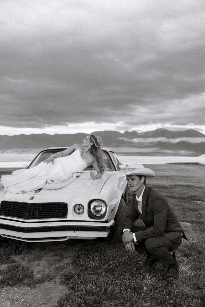 Black and white photo of bride draped across hood of vintage Camaro while groom in cowboy hat crouches beside it under stormy Montana sky — Micah + Greg — Ember & Altar Wedding — Polson Montana — Stacy Edwards Photography