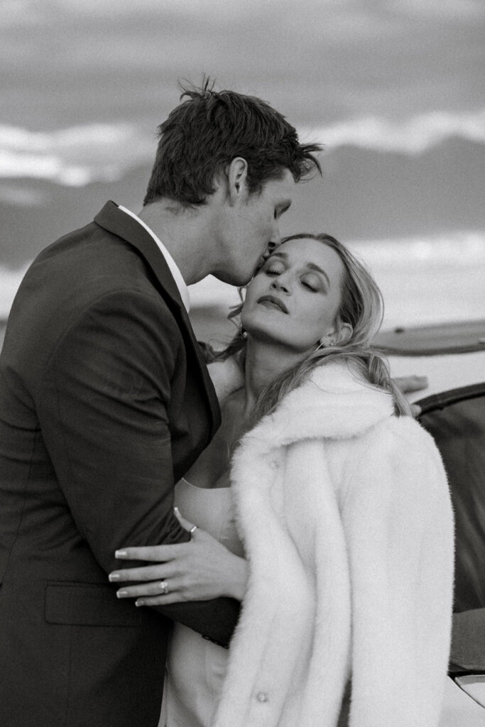 Black and white close-up of groom kissing bride on the forehead while she leans into him wearing a white fur wrap — Micah + Greg — Ember & Altar Wedding — Polson Montana — Stacy Edwards Photography