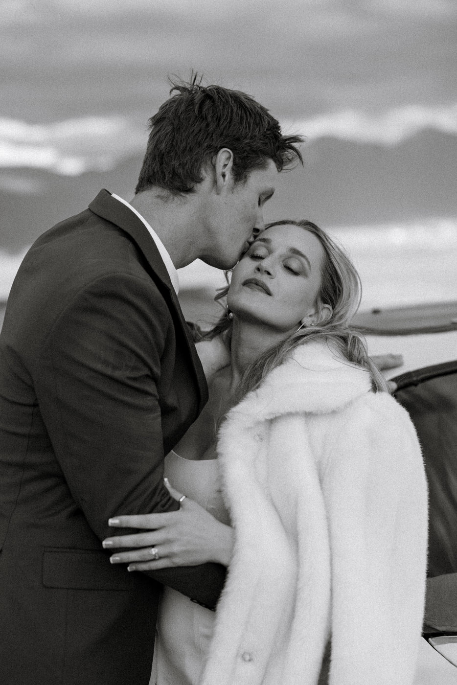 Black and white close-up of groom kissing bride on the forehead while she leans into him wearing a white fur wrap — Micah + Greg — Ember & Altar Wedding — Polson Montana — Stacy Edwards Photography