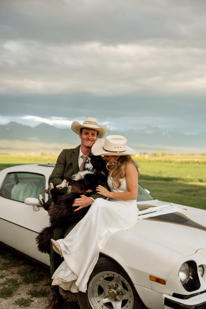 Bride and groom in matching cowboy hats with their black and white dog on the hood of a vintage Camaro with Mission Mountains behind them — Micah + Greg — Ember & Altar Wedding — Polson Montana — Stacy Edwards Photography