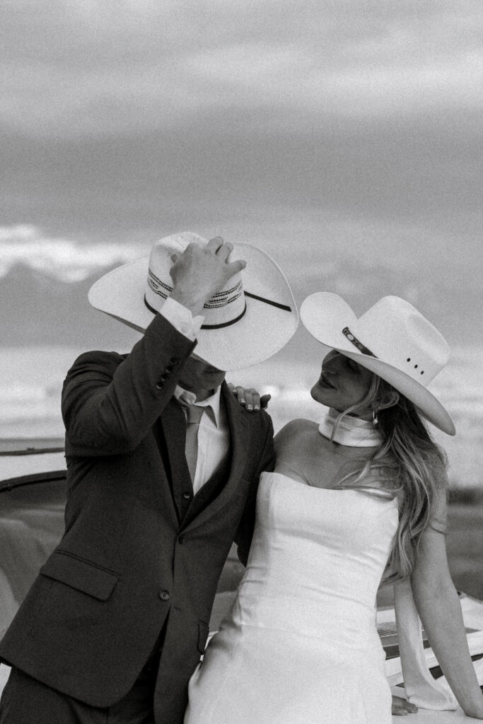 Black and white photo of bride and groom both in cowboy hats laughing together beside the vintage Camaro under stormy skies — Micah + Greg — Ember & Altar Wedding — Polson Montana — Stacy Edwards Photography