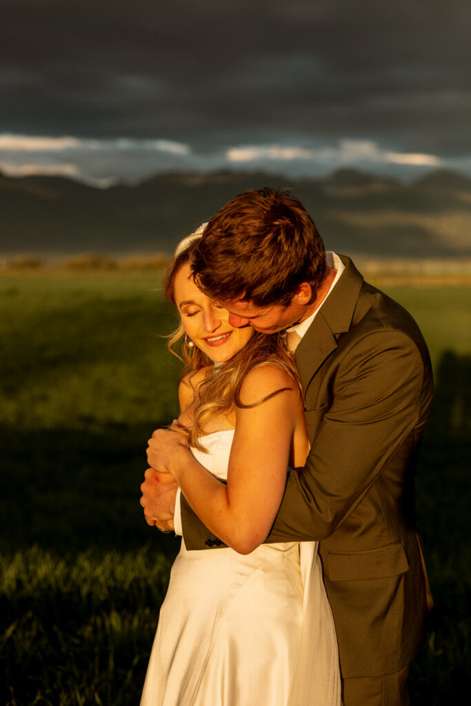Groom holding bride from behind and kissing her cheek in dramatic golden evening light with dark storm clouds and Mission Mountains behind them — Micah + Greg — Ember & Altar Wedding — Polson Montana — Stacy Edwards Photography