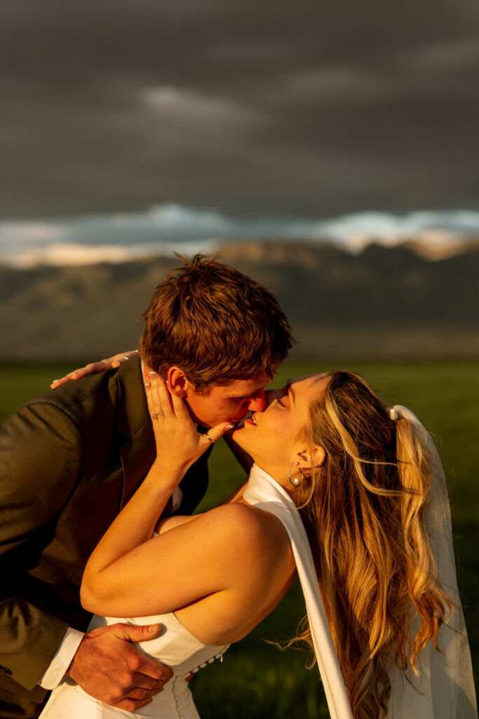 Close-up of bride and groom kissing at golden hour with Mission Mountains and dramatic storm clouds behind them in open Montana field — Micah + Greg — Ember & Altar Wedding — Polson Montana — Stacy Edwards Photography