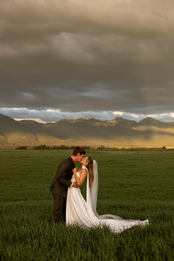 Bride and groom kissing in open green field at stormy sunset with the Mission Mountains glowing behind them and her veil trailing in the wind — Micah + Greg — Ember & Altar Wedding — Polson Montana — Stacy Edwards Photography