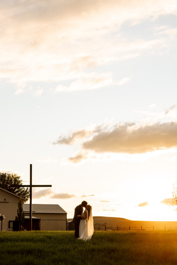 Bride and groom silhouetted kissing at golden sunset beside a wooden cross at Ember and Altar — Micah + Greg — Ember & Altar Wedding — Polson Montana — Stacy Edwards Photography