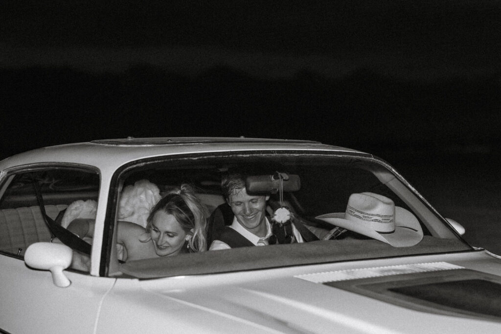 Black and white photo of bride leaning on groom's shoulder as they laugh together inside a vintage Camaro at the end of the night — Micah + Greg — Ember & Altar Wedding — Polson Montana — Stacy Edwards Photography