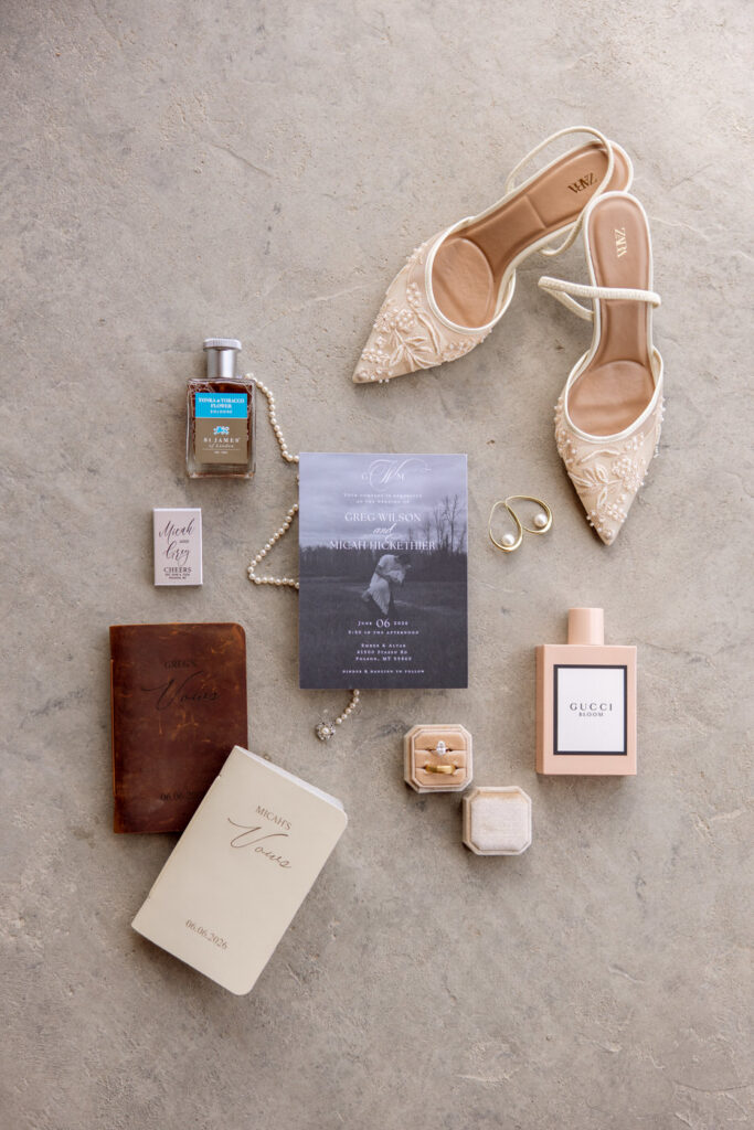 Wedding detail flatlay with invitation suite, vow books, rings, bridal shoes, pearl necklace, and perfume — Micah + Greg — Ember & Altar Wedding — Polson Montana — Stacy Edwards Photography