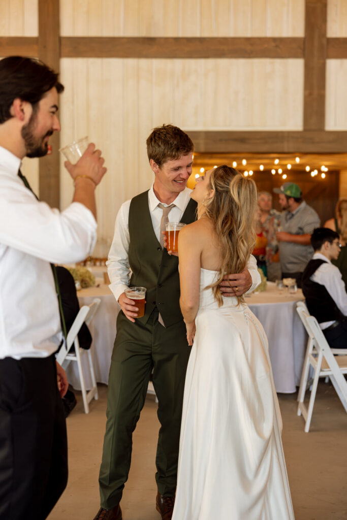 Bride and groom holding drinks and smiling at each other during reception inside the Ember and Altar barn — Micah + Greg — Ember & Altar Wedding — Polson Montana — Stacy Edwards Photography