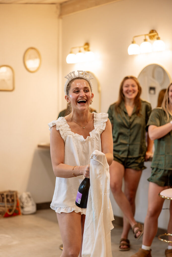 Bride laughing and holding champagne bottle while getting ready with bridesmaids in green pajama sets — Micah + Greg — Ember & Altar Wedding — Polson Montana — Stacy Edwards Photography