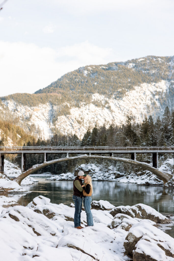 Engaged couple sharing a kiss on the snowy banks of the Middle Fork Flathead River below Belton Bridge — Lindsey and Ethan — West Glacier Glacier National Park Engagement Photographer — Stacy Edwards Photography
