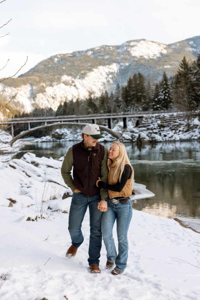 Engaged couple walking hand in hand and laughing along the snowy riverbank below Belton Bridge near West Glacier — Lindsey and Ethan — Glacier National Park Winter Engagement Session — Stacy Edwards Photography